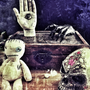 A grunge style picture of a Palmistry hand, a zombie hand, a voodoo doll and an ornate skull on an old wooden box.
