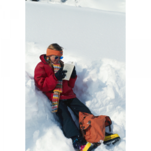 A picture of a snow boarder sat in the snow reading a book