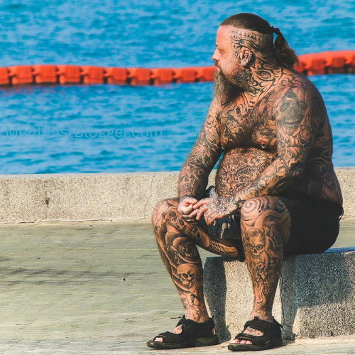 A picture of a heavily tattooed man sat next to the sea.